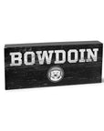 Grey wooden block with distressed imprint of BOWDOIN in white over a black, white and grey mascot medallion.