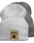 Two ribbed knit beanie hats shown on a white background, with a light heather gray one in front and a darker gray one behind. The front hat has a tan rectangular patch with the word "BOWDOIN" and a paw print.