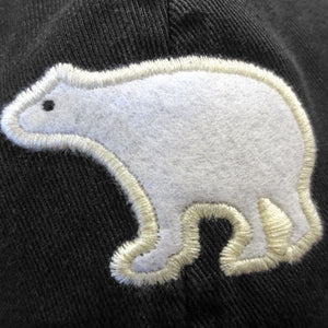 A closeup shot of the white felt polar bear embroidered in white on a black twill hat. The polar bear is in profile and has a small eye embroidered in black on its face.
