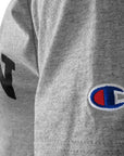 Closeup shot of the left sleeve of a short-sleeved Oxford heather gray T-shirt showing an embroidered Champion C logo patch in red, white, and blue.