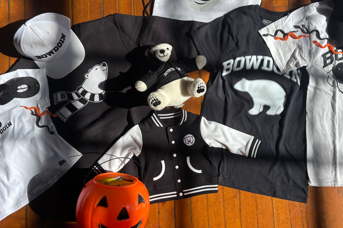 A flat-lay arrangement of Bowdoin branded merchandise on a wooden floor, including black and white t-shirts, a varsity jacket, a white cap, a teddy bear, and a jack-o'-lantern bucket.