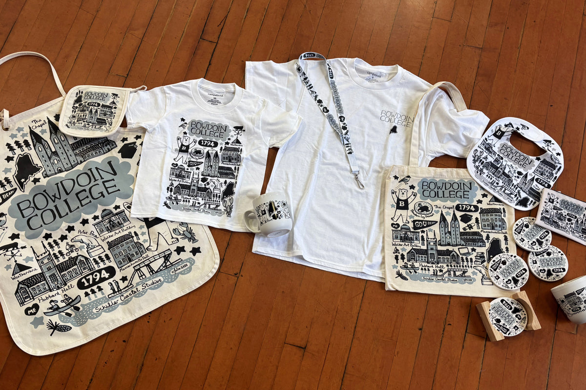 Flat-lay arrangement of Julia Gash for Bowdoin merchandise including T-shirts, coasters, mugs, a bag, a bib, and an apron. All items are styled on a warm wooden floor.