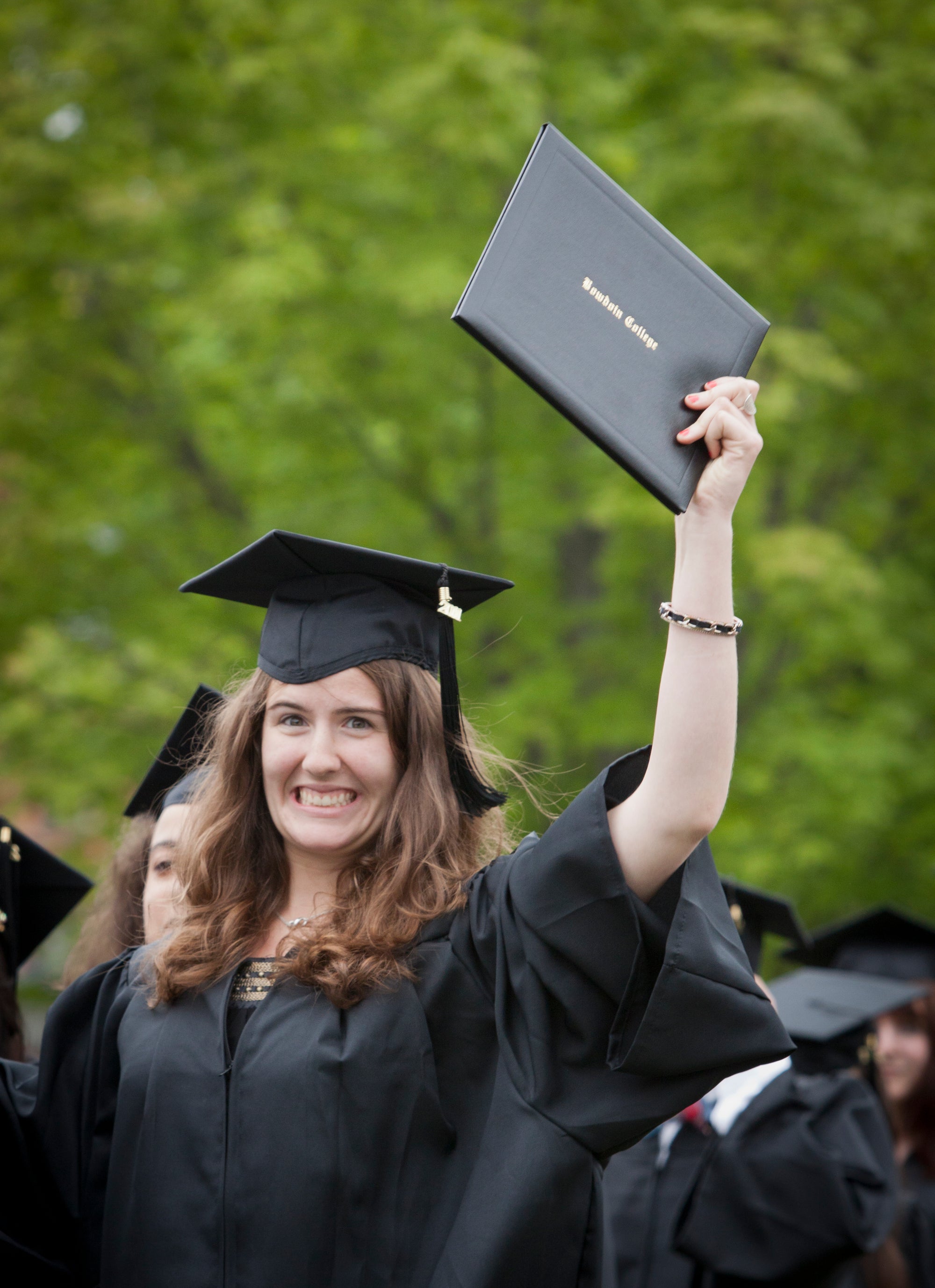 Graduate in cap and gown holding a Bowdoin diploma with greenery in the background