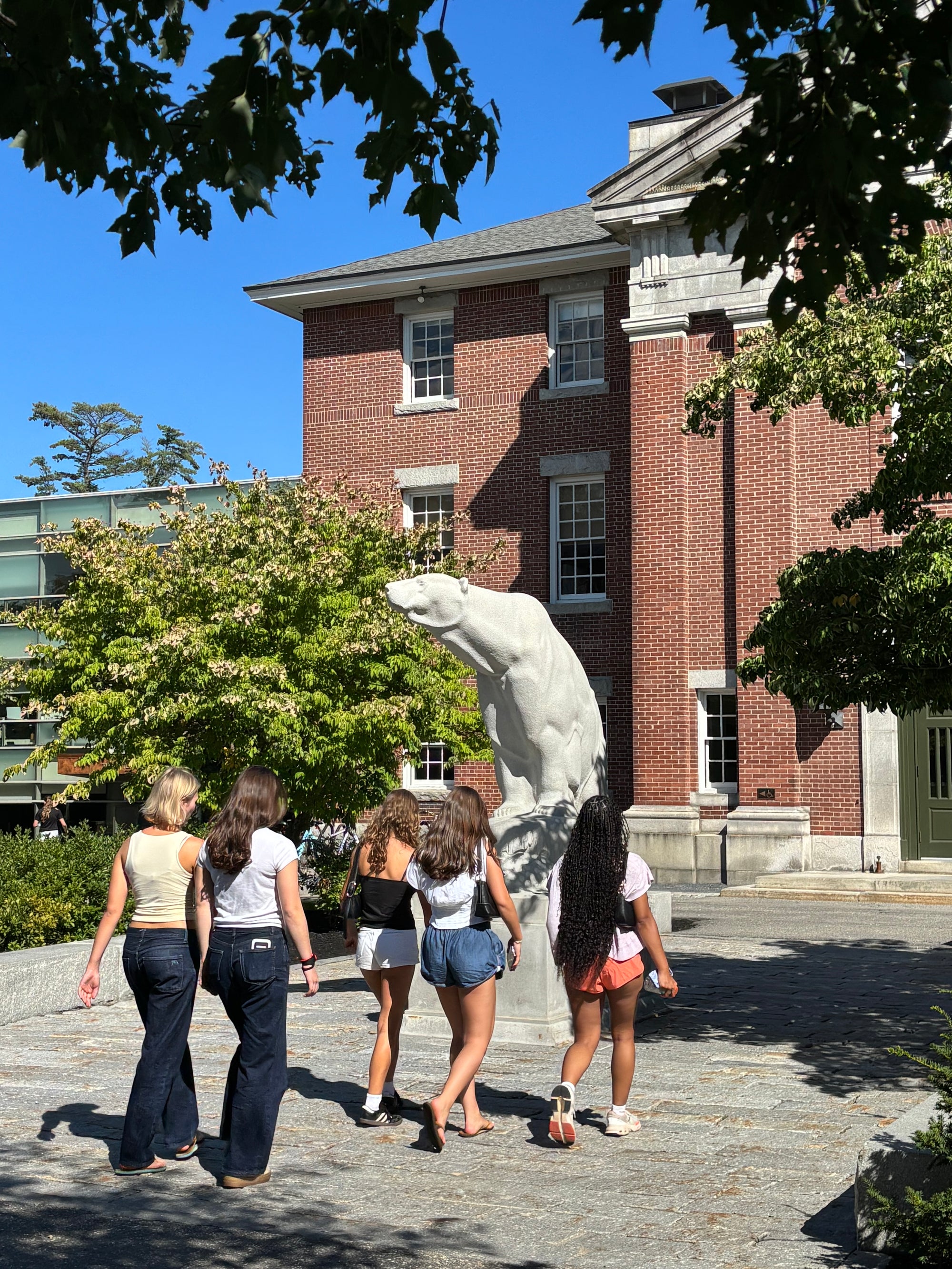 Five women walking away from the camera in front of a large white polar bear sculpture on a stone plaza, with a red brick building and green trees in the background under a blue sky.
