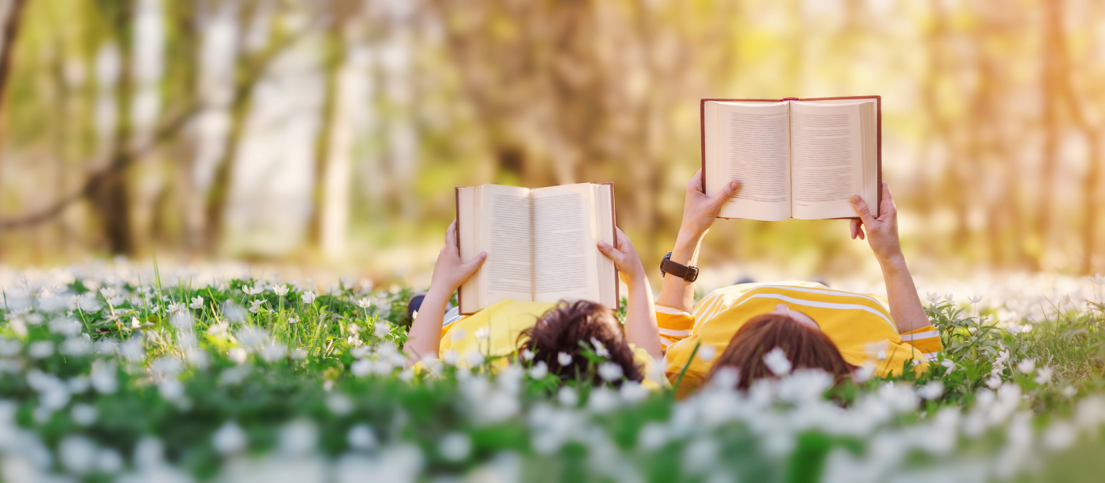 Two people lying on grass reading books in a park.