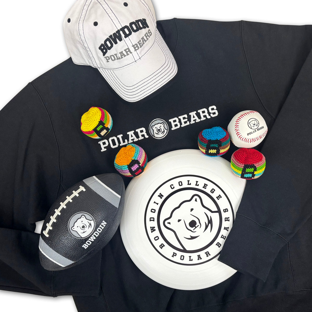 A flat-lay arrangement of Bowdoin Polar Bears themed merchandise on a white background, including a black sweatshirt with white text, a white baseball cap, a white frisbee, a black football, and several colorful knitted footbags.