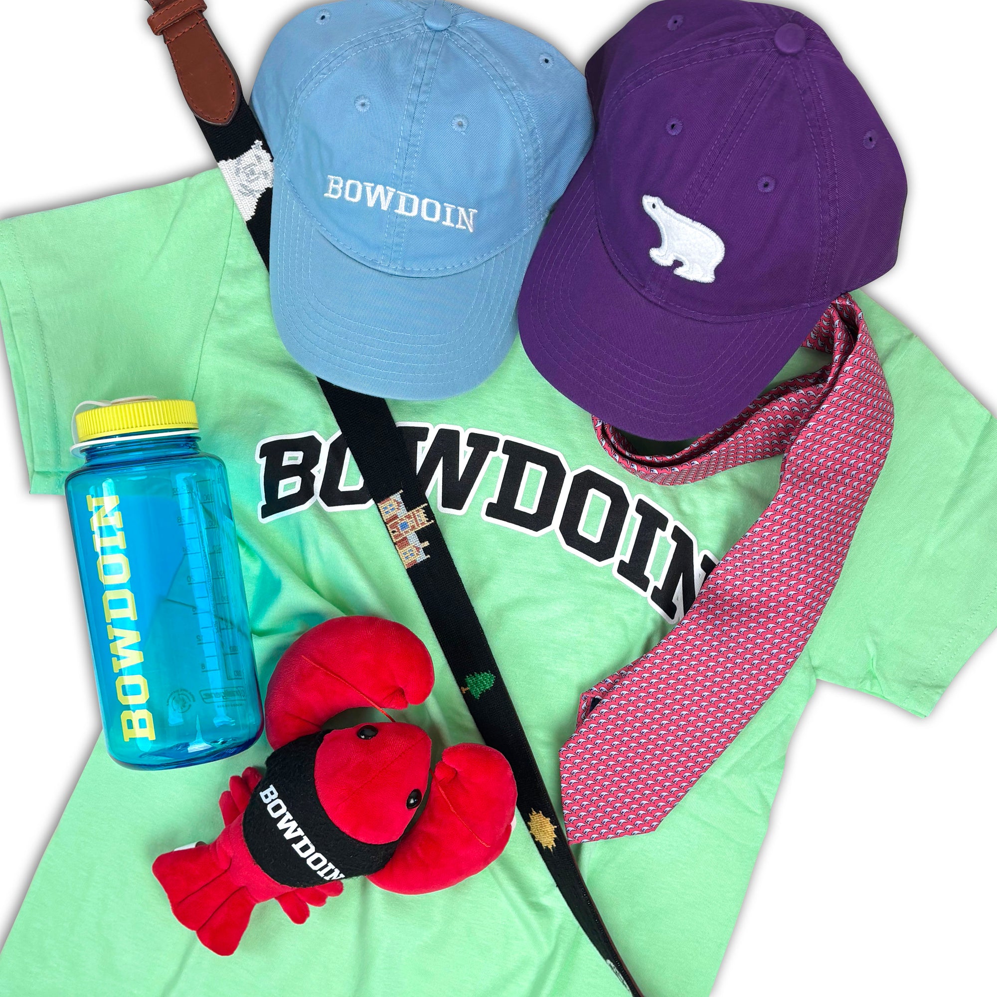 A flat-lay arrangement of Bowdoin-themed merchandise on a white background, including a mint green t-shirt with 'BOWDOIN' printed across the chest, a light blue cap and a purple cap with white Bowdoin logos, a blue water bottle, a pink patterned necktie, a red lobster plush toy, and a black belt with colorful embroidery.