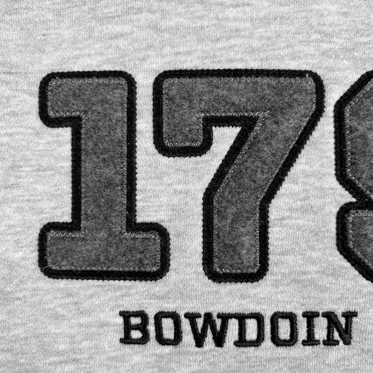 Close-up of a light gray heathered fabric with large dark gray embroidered numbers '179' and the word 'BOWDOIN' in black lettering below.