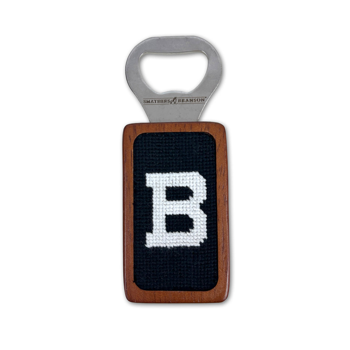 A Smathers &amp; Branson bottle opener with a rectangular wooden body and a black needlepoint center featuring a white embroidered letter 'B'.