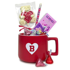 A red mug with a square handle and a white imprint of a heart with a Bowdoin B cutout. The mug contains 5 Hershey's kisses is pink, red, and silver foil, a pink box of Brach's tiny conversation hearts, a purple pencil covered with pink hearts, and a white chocolate lollipop in the shape of a white teddy bear holding a red heart.