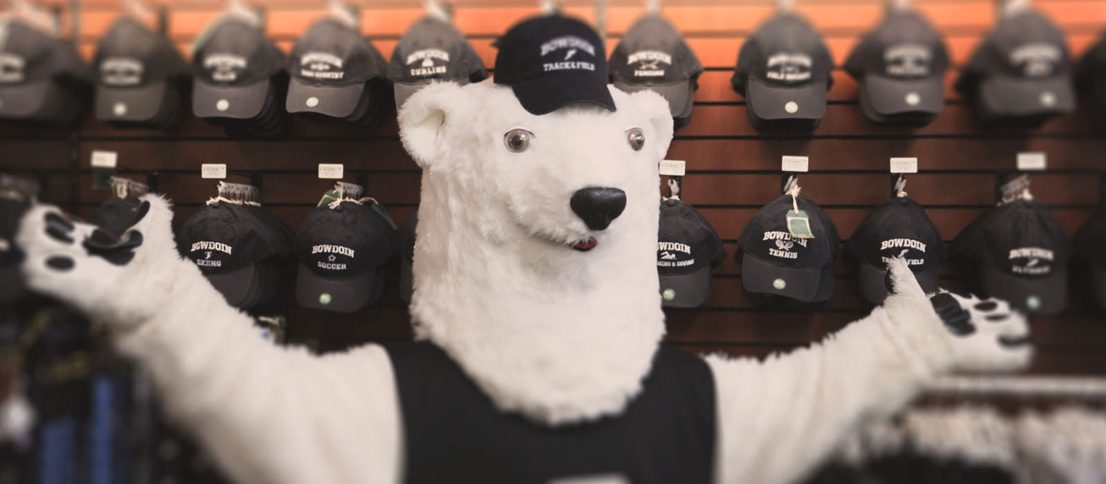 Polar bear mascot wearing Bowdoin hat in front of wall of Bowdoin hats in the SU Bowdoin Store.