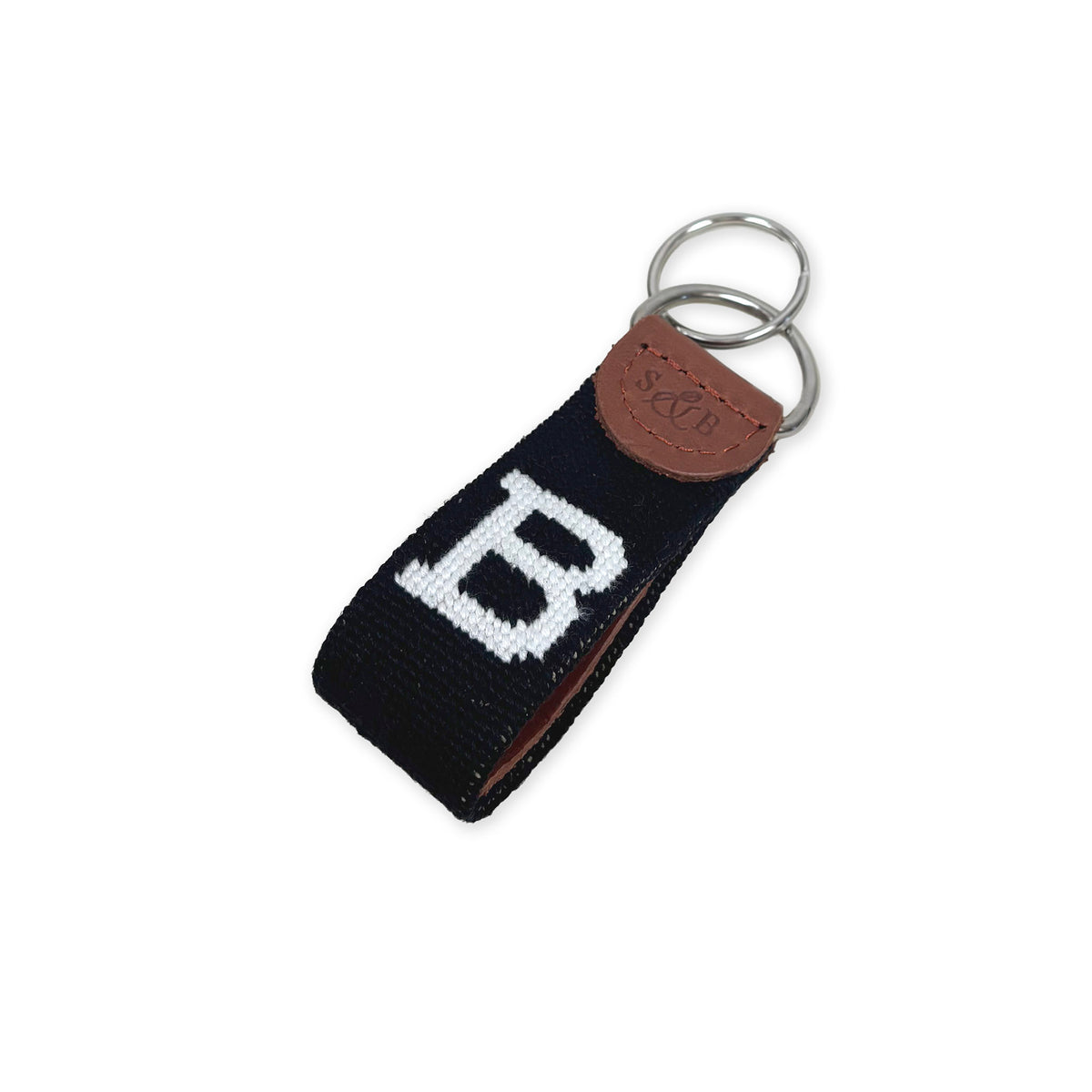 A black needlepoint key fob with a large white letter 'B' and a brown leather patch embossed with 'S&amp;B', attached to steel key rings on a white background.