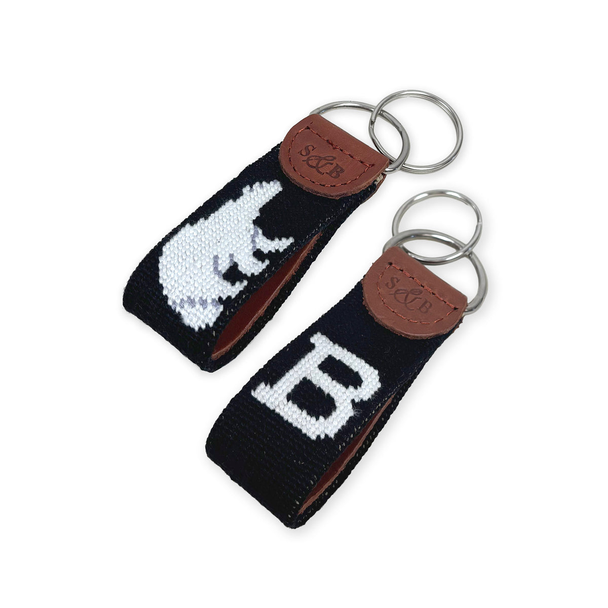 Two black needlepoint key fobs with brown leather tabs and silver key rings, each featuring a white embroidered letter or animal motif, shown on a white background.
