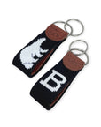 Two black needlepoint key fobs with brown leather tabs and silver key rings, each featuring a white embroidered letter or animal motif, shown on a white background.